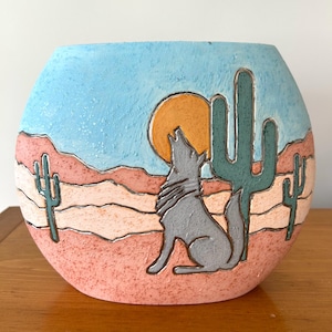 Könnte beinhalten: Ein runder, handbemalter Keramiktopf mit einer Wüstenlandschaft. Der Topf zeigt einen heulenden Wolf, eine untergehende Sonne, Kakteen und geschichtete Berge in Blau-, Orange-, Rosa- und Grüntönen. Der Topf ist ein dekorativer Gegenstand.