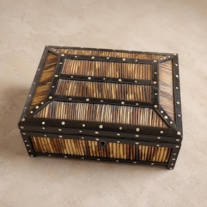 Peut inclure: Une boîte de rangement rectangulaire dotée d'un couvercle et de côtés composés de fines lattes verticales rayées marron et beige. La boîte est encadrée par des bordures marron foncé incrustées de petits points circulaires blanc cassé.