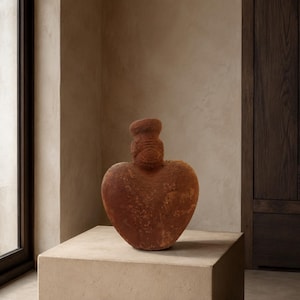 May include: A heart-shaped terracotta vase with a textured, earthy surface and a narrow, cylindrical neck. The vessel features a reddish-brown color and a porous, matte finish, showcasing a distinct, rounded silhouette that tapers toward the top.