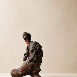 Peut inclure: Sculpture abstraite d'une figure assise, d'aspect vieilli. L'œuvre est réalisée dans un matériau sombre et texturé, peut-être du métal ou de la pierre, avec un aspect rugueux et vieilli. La sculpture est montée sur un petit socle noir.