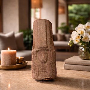 Könnte beinhalten: Eine verwitterte, rechteckige, braune Steinskulptur einer Figur mit hohem Kopfschmuck. Die Skulptur steht auf einem hellfarbigen Tisch. Eine brennende Kerze und eine Vase mit weißen Blumen befinden sich im Hintergrund.