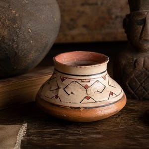 Peut inclure: Un petit pot en céramique à profil bas avec une base large et arrondie et un bord évasé. L'extérieur présente une section supérieure de couleur crème décorée de motifs géométriques noirs et de petits losanges rouges, sur une base en terre cuite.