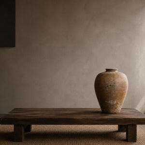 Puede incluir: Una vasija de cerámica grande y desgastada de color beige, con un cuerpo ancho y un cuello estrecho, se asienta sobre una mesa baja rectangular de madera marrón oscuro. La vasija presenta una superficie mate y texturizada con marcas.