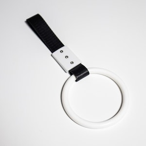 May include: A white circular handle with a black strap and a white rectangular base. The handle is connected to the strap with a white rectangular base that has three small silver rivets. The background is white.