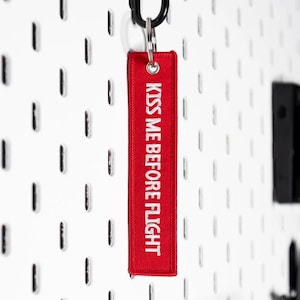 May include: A red rectangular keychain tag with the embroidered text "KISS ME BEFORE FLIGHT" in white. The tag is attached to a black metal ring. The background is a white pegboard.