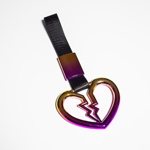 May include: A metallic keychain featuring a broken heart design. The heart transitions from gold to purple, with a black strap and a matching gradient metal clasp. The keychain is a symbolic accessory.