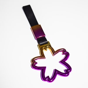 May include: A metallic flower-shaped keychain with a gradient finish, transitioning from gold to purple. It features a black strap and a rectangular connector. The design is modern and eye-catching.