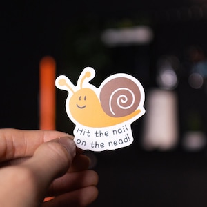 May include: A sticker depicting a cartoon snail with a yellow body and a brown shell. The snail has a smiling face and the text "Hit the nail on the nead!" in a playful font. The sticker is held by a hand.
