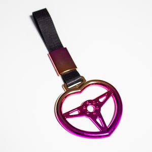 May include: A heart-shaped keychain with a steering wheel design. The keychain features a gradient of colors, transitioning from gold to pink and purple. It has a black strap and a metal clasp.