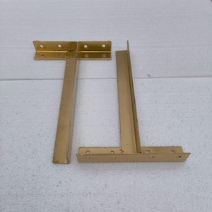 Op de afbeelding: Twee gouden metalen tafelpoten met een T-vormig ontwerp. Elk been heeft een rechthoekige basis en een verticale steun.