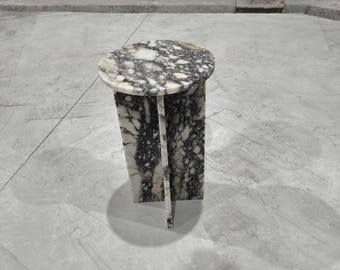 May include: A round marble side table with a white and gray marbled top and a white base with gray marbling. The table is standing on a gray concrete floor.