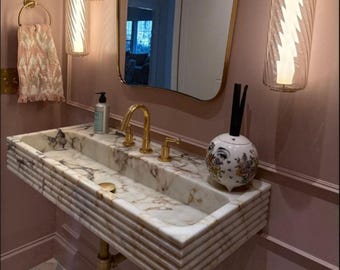Può includere: Un lavabo rettangolare a parete in marmo con venature marroni e un design esterno a coste. È dotato di un rubinetto dorato, uno scarico dorato abbinato e un diffusore in ceramica decorativo con una stampa floreale e un volto.