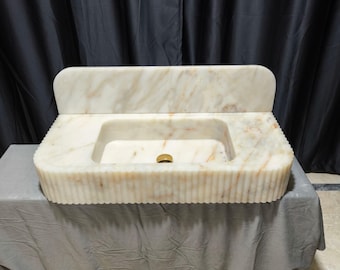 May include: A white marble bathroom sink with a backsplash and a fluted design. The sink has a single drain hole and a gold-colored drain.