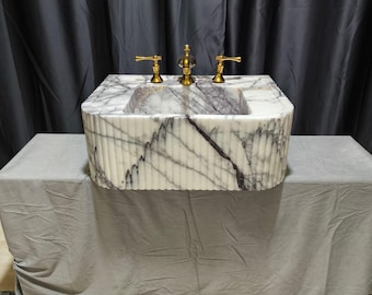 May include: A white marble sink with a rippled texture and purple veins. The sink has three gold taps and is mounted on a grey countertop.