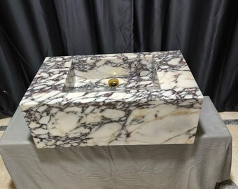 May include: A white marble sink with purple and brown veins. The sink is rectangular and has a square basin. The sink is sitting on a gray surface.