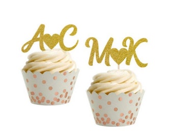 May include: Two gold glitter cupcake toppers with hearts and initials "A & C" and "M & K". The toppers are on cupcakes with white frosting and white cupcake liners with rose gold polka dots.