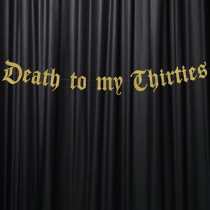 May include: A gold banner with the phrase "Death to my Thirties" in an ornate font hangs against a black curtain backdrop. The banner is strung on a thin, nearly invisible string, creating a floating effect.