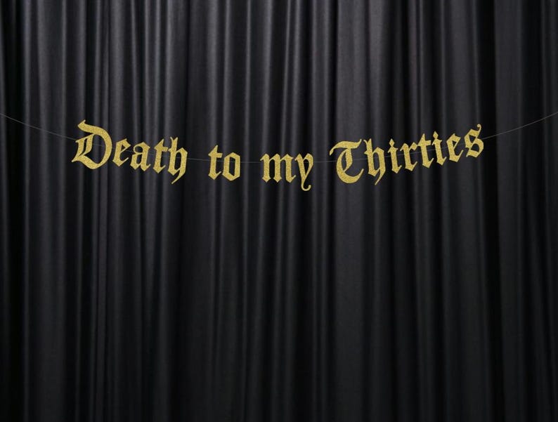 May include: A gold banner with the phrase "Death to my Thirties" in an ornate font hangs against a black curtain backdrop. The banner is strung on a thin, nearly invisible string, creating a floating effect.