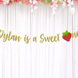 Op de afbeelding: Gouden glitterbanner met de tekst "Dylan is a Sweet One" en een rode aardbeienafbeelding.