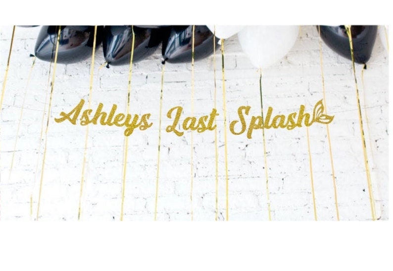 May include: Gold glitter letters spelling "Ashley's Last Splash" with a mermaid tail symbol hanging from white string against a white brick wall background.