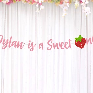Op de afbeelding: Roze glitterbanner met de tekst "Dylan is a Sweet One" en een rode aardbeienafbeelding.