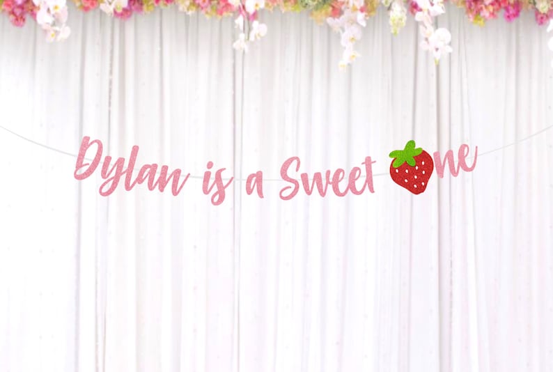 Op de afbeelding: Roze glitterbanner met de tekst "Dylan is a Sweet One" en een rode aardbeienafbeelding.