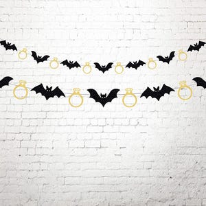 May include: A decorative banner consisting of two horizontal strings featuring alternating black glitter bat silhouettes and gold glitter engagement ring shapes. The cutouts are evenly spaced along thin white strings, creating a repeating pattern.
