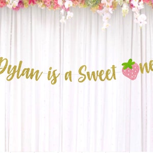 Op de afbeelding: Gouden glitterbanner met de tekst "Dylan is a sweet one" en een roze aardbeienafbeelding.
