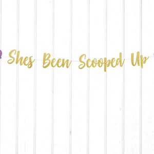 May include: Gold glitter banner with the text "She's Been Scooped Up" and two purple glitter ice cream cones on either end.