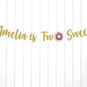 Puede incluir: Pancarta de purpurina dorada con el texto "Amelia is Two Sweet" y un gr&aacute;fico de donut morado y blanco.