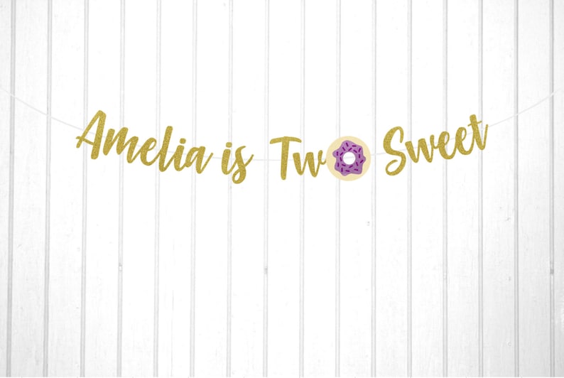 Puede incluir: Pancarta de purpurina dorada con el texto "Amelia is Two Sweet" y un gr&aacute;fico de donut morado y blanco.
