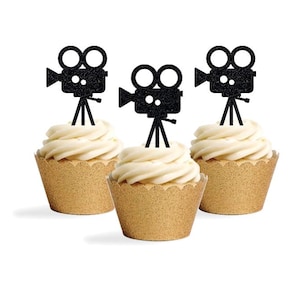 May include: Three cupcakes with white frosting and gold cupcake wrappers. Each cupcake is topped with a black glitter film camera-shaped decoration. The cupcakes are arranged on a white background.