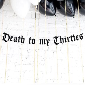 May include: A banner with the phrase "Death to my Thirties" in black gothic lettering hangs against a white brick wall. Black and white balloons are visible at the top, with gold string.