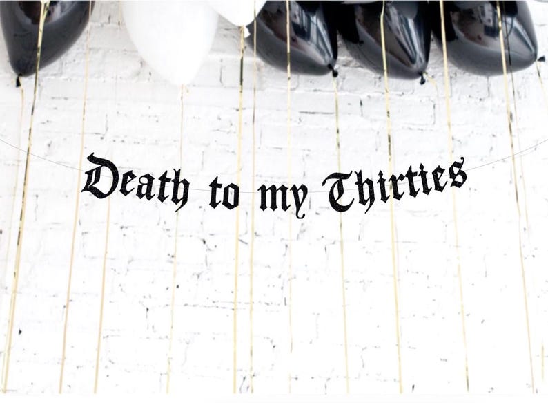 May include: A banner with the phrase "Death to my Thirties" in black gothic lettering hangs against a white brick wall. Black and white balloons are visible at the top, with gold string.