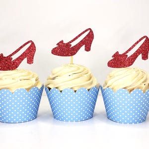 May include: Three cupcakes with white frosting and red glitter shoe toppers. The cupcakes are in blue polka dot liners. The toppers are shaped like high-heeled shoes.