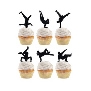 May include: Six cupcakes with black silhouette toppers of people doing breakdancing moves. The cupcakes are frosted with white frosting.