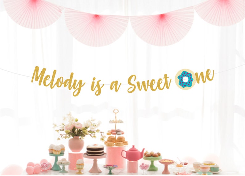 May include: Gold glitter banner with the text "Melody is a sweet one" and a blue and pink donut graphic. The banner is hanging from a white ceiling with pink paper fans.