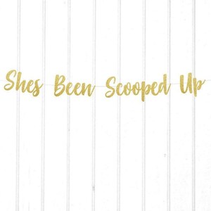 Puede incluir: Pancarta de purpurina dorada con el texto "She's Been Scooped Up" y dos recortes de conos de helado, uno rosa y otro verde.