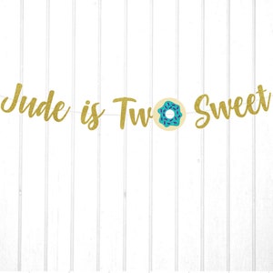 Puede incluir: Pancarta de brillo dorado con el texto "Jude is Two Sweet" y un gr&aacute;fico de donut azul y blanco.