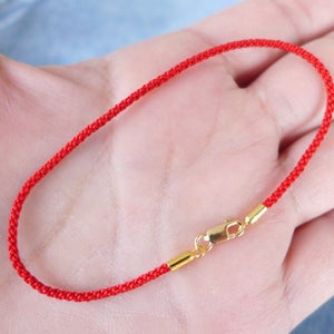 May include: A red braided bracelet with a gold-colored clasp and end pieces. The bracelet is made of a thin, woven cord and is designed to be worn on the wrist. The clasp is a simple hook and eye closure.