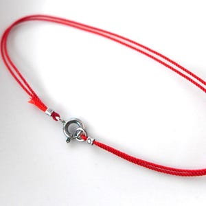 May include: A thin red cord bracelet or necklace featuring a double-strand design. The jewelry piece is secured with a circular silver-toned spring ring clasp and small metal crimp beads at the connection points, creating a simple, minimalist look.