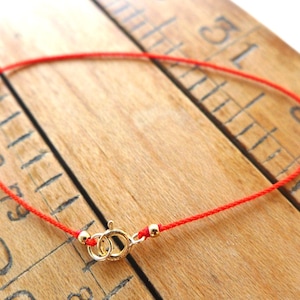 May include: A red string bracelet with gold-colored clasp and accents. The bracelet is displayed on a wooden surface with measurement markings. The bracelet is a simple design, suitable for everyday wear.