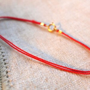 May include: A delicate red cord bracelet with a gold-colored clasp. The bracelet is made of two thin strands of red cord. The clasp is a small, round design. The bracelet is displayed on a beige fabric surface.