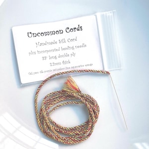 May include: A spool of handmade silk cord with an incorporated beading needle. The cord is 33 inches long, double ply, and 2.2mm thick. The cord is a mix of yellow, red, and purple colors. The text "Uncommon Cords" is visible.
