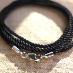 May include: Black rope necklace with a silver clasp. The necklace is made of tightly woven black rope, forming a thick, textured cord. The clasp and end caps are silver-colored metal. The necklace is displayed on a neutral-colored fabric.