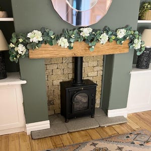 May include: Miniature fireplace scene with a black stove, brick chimney, and wooden mantel decorated with white flowers and greenery. A round mirror hangs above the mantel, and a patterned rug sits on the floor.