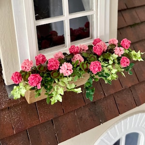 May include: A miniature window box filled with bright pink and pale pink flowers and green leaves. The window box is light brown and rests on a brown tiled roof, beside a white window frame.