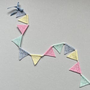 May include: A pastel-colored fabric bunting with a blue ribbon. The bunting is made of alternating light blue, yellow, pink, and green triangles with a white floral pattern.