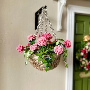 May include: A miniature hanging basket filled with pink flowers and green foliage. The basket is made of woven material and hangs from a silver chain attached to a black bracket. The flowers are a vibrant pink, adding a pop of color.