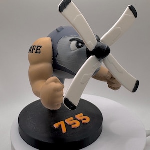 Puede incluir: Figura impresa en 3D de un personaje musculoso con cabeza de hélice. La figura tiene un casco gris, brazos color canela y una base negra con el número "755" en naranja. Las aspas de la hélice son blancas con puntas negras.